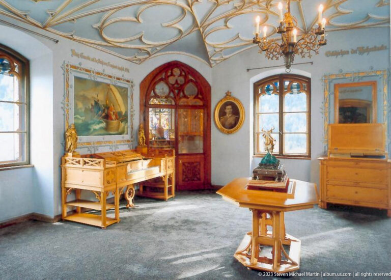 Light-blue Hohenschwangau chamber with painted ceiling, red-trimmed arched doorway, tables, chairs, and cabinets.