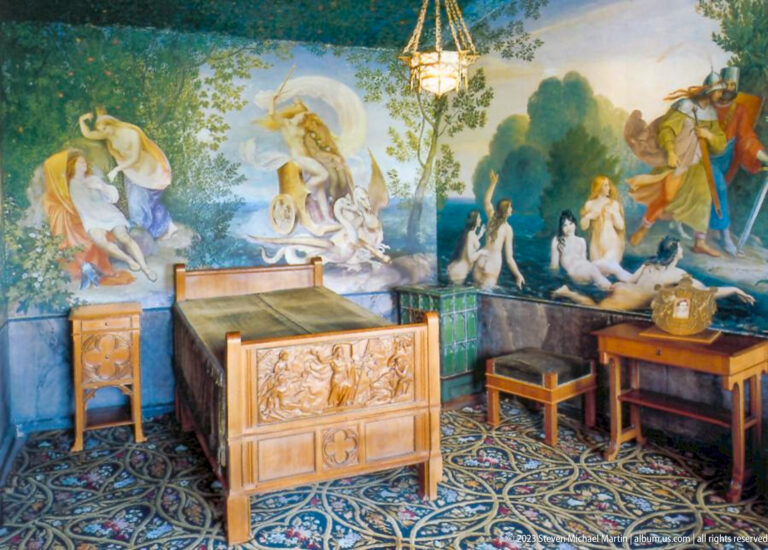 Carved bed beneath large mythological wall paintings, with patterned floor and small furniture in Hohenschwangau Castle.