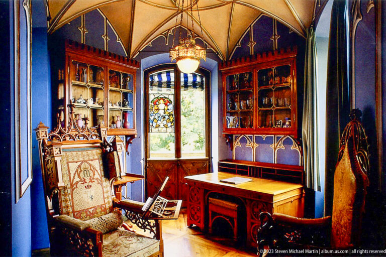 Dark blue study with carved wood shelves, writing desk, upholstered chair, and stained-glass window at Hohenschwangau Castle.