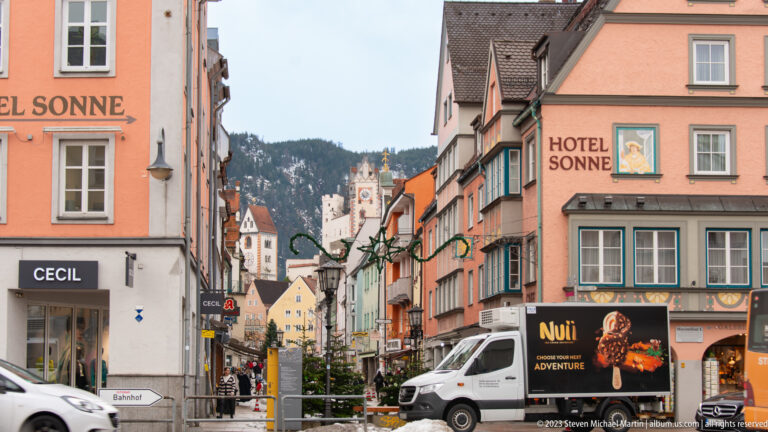 Pastel buildings and shops line a Füssen street, with a historic tower and wooded mountain slope beyond.