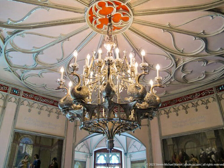 Ornate metal chandelier with sculptural animal forms hanging beneath a painted ribbed ceiling and red floral medallion.