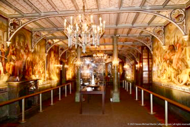 Long gallery with wall-to-wall murals, patterned ceiling, chandeliers, columns, and a central visitor path.