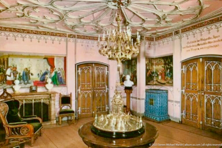 Pink historic room with chandelier, murals, carved doors, blue ceramic stove, upholstered chair, and round display table.