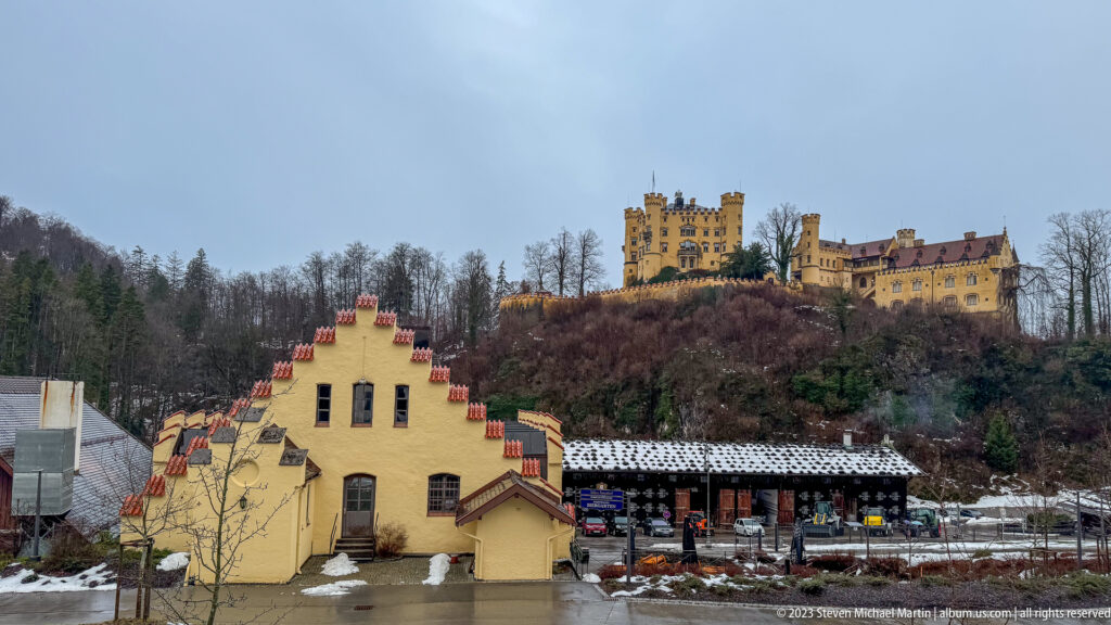 Ochre Hohenschwangau Castle on a wooded ridge above pale village buildings and snow patches.