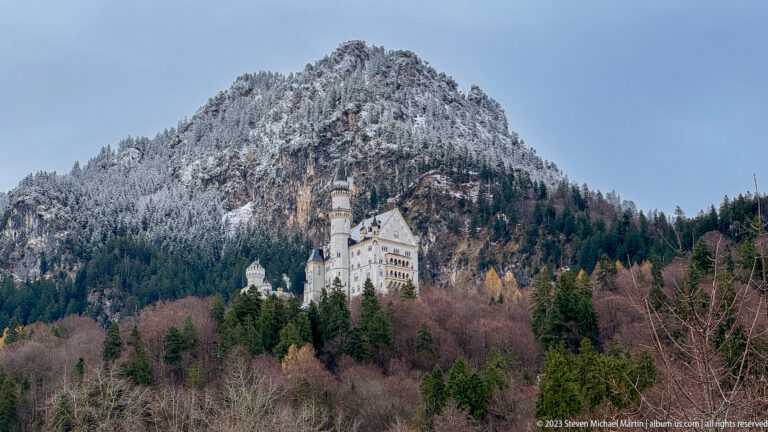Neuschwanstein Castle on a wooded slope below a steep snow-dusted mountain and dark conifer forest.