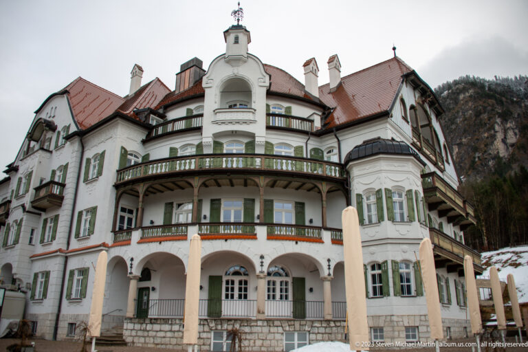 Large white hotel-style building with curved balconies, bay windows, green shutters, arches, and red roofs.