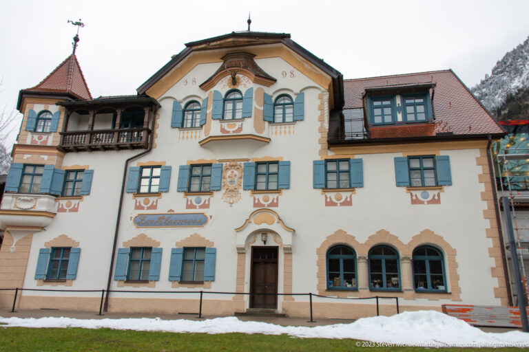 Cream lodging facade with blue shutters, painted ornament, central gable, decorative windows, and snow at ground level.
