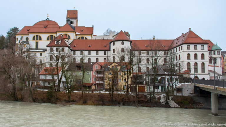Pale Baroque buildings of St. Mang Abbey rise beside the Lech, with red roofs and a bridge at right.