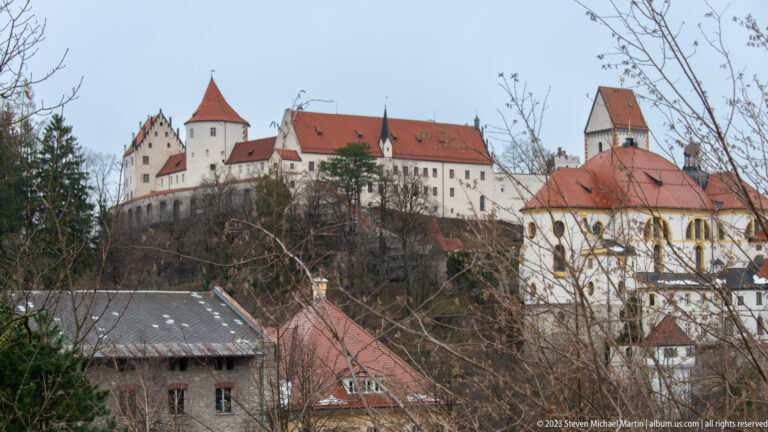 High Castle of Füssen on a hill with red roofs and towers, seen beyond bare branches and nearby historic buildings.