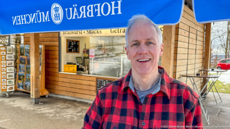 Smiling man in a red-and-black plaid jacket standing outside a small wooden snack kiosk.