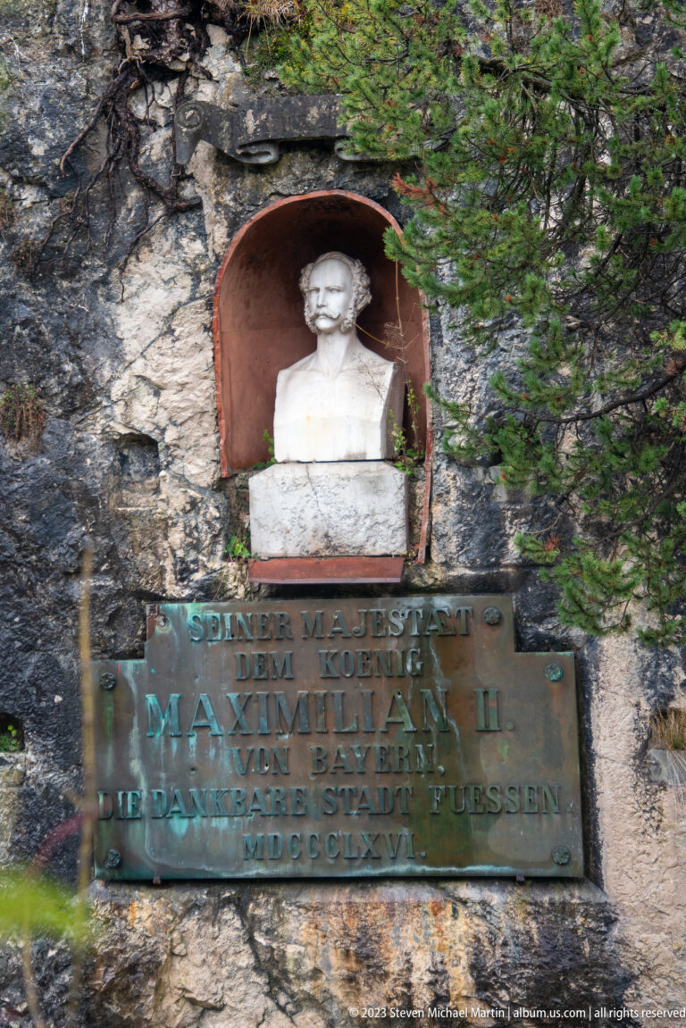 White bust of Maximilian II in a rock niche above a bronze German inscription plaque.