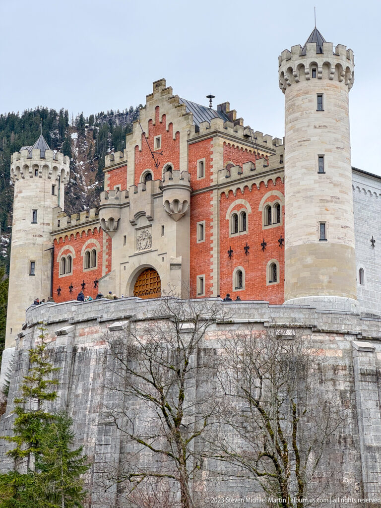 Red Neuschwanstein gatehouse and pale crenellated towers rise above a massive stone retaining wall.