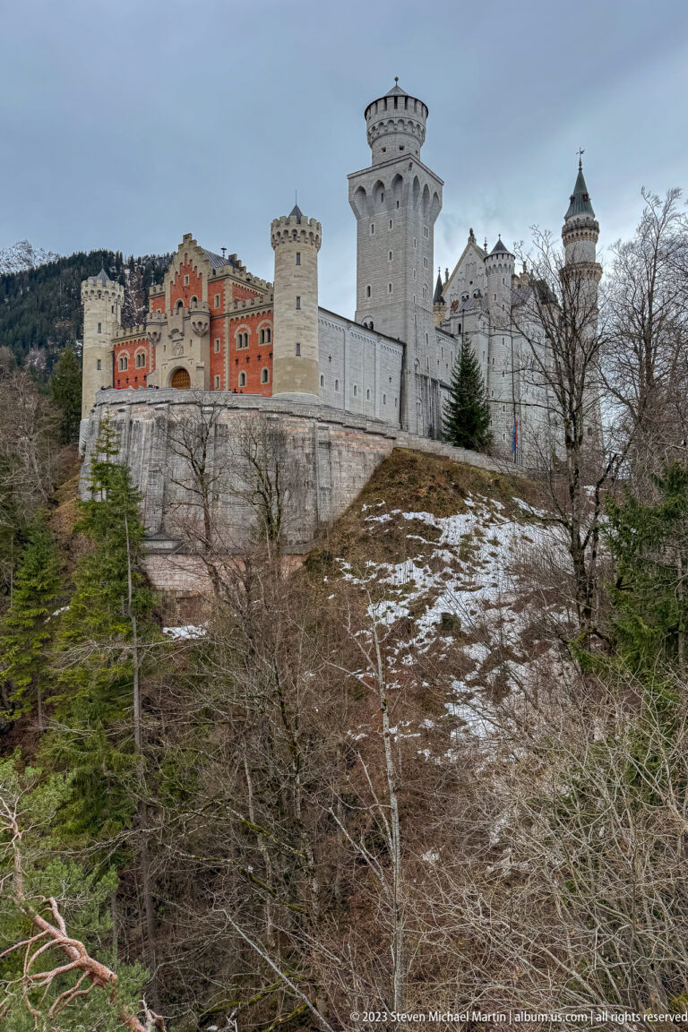 Neuschwanstein Castle stretches across a rocky, partly snow-covered hill above conifer forest.