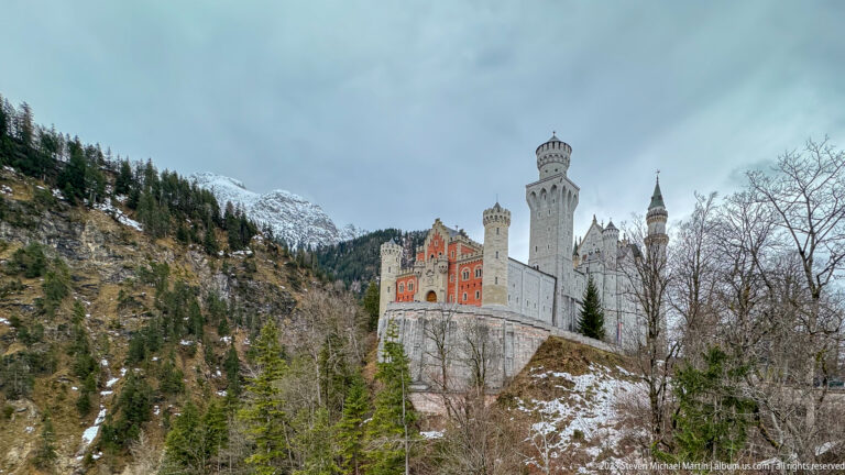 Neuschwanstein Castle rises above forest with snow-streaked Alpine slopes behind its red and white towers.