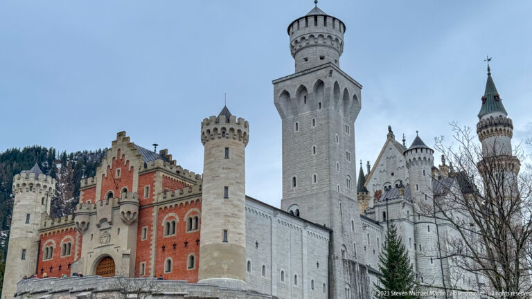 Neuschwanstein’s red gatehouse, pale towers, and tall square tower fill the frame against a gray sky.