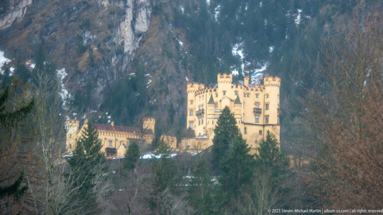 Yellow Hohenschwangau Castle with crenellated towers stands on a wooded mountainside below rocky cliffs.