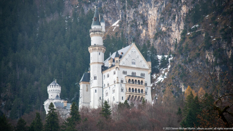 White Palas and towers of Neuschwanstein Castle stand against dark forest and a rocky mountain face.