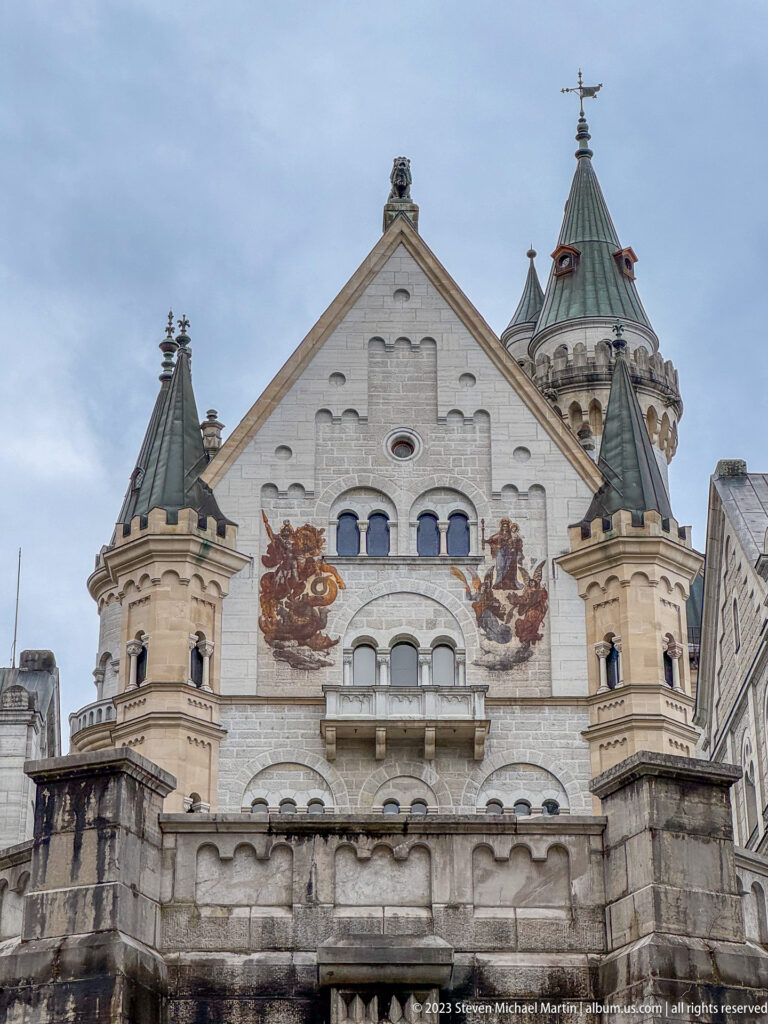 Pale Neuschwanstein gable with arched windows, flanking turrets, and large painted heraldic lions.