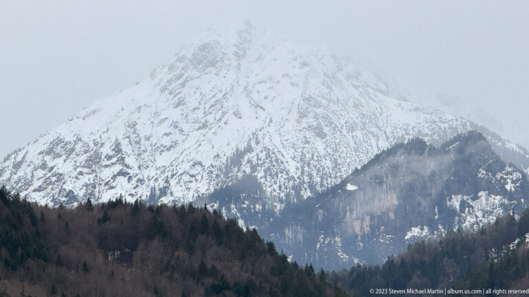 Steep snow-covered Alpine peak partly obscured by low cloud above dark forest.