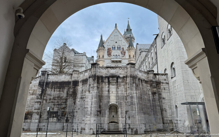 Stone arch frames Neuschwanstein’s ornate gable and towers above a dark weathered courtyard wall.