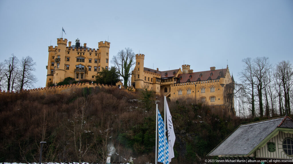 Hohenschwangau Castle above a wooded winter hillside, with ochre towers and long residential wings beneath a gray sky.