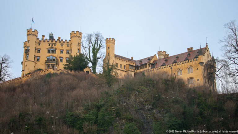 Hohenschwangau Castle extending across a wooded ridge, with crenellated towers and ochre walls visible through leafless trees.