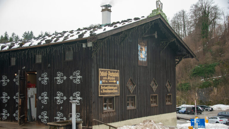 Dark timber building with white painted ornament, Bavarian shield, hanging sign, and snow along the roof and ground.