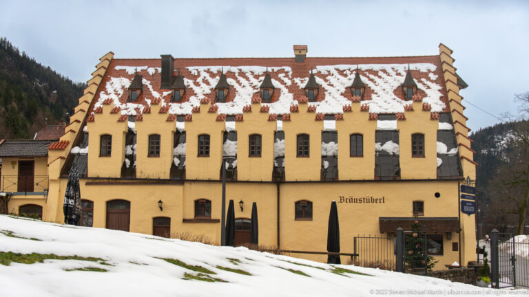 Yellow Bräustüberl building with stepped gables, rows of dormer windows, and patches of snow on the steep roof.