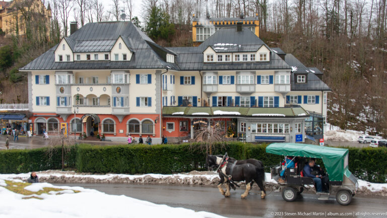 Large pale hotel-style complex with blue shutters and steep roofs behind a horse-drawn carriage on a snowy roadside.