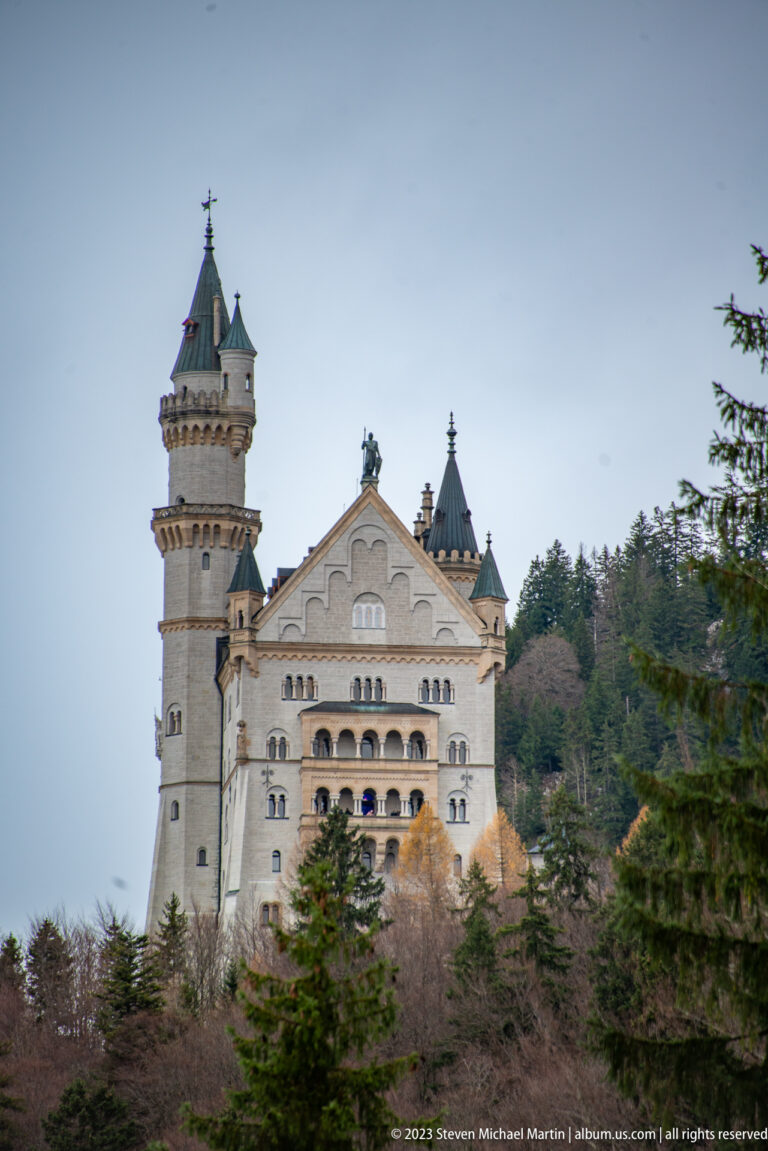 Neuschwanstein Castle rising above trees, with a tall cylindrical tower and pale palace façade against an overcast sky.