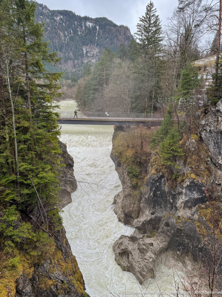 Fast pale river water runs through a rocky gorge beneath a low bridge near Füssen.