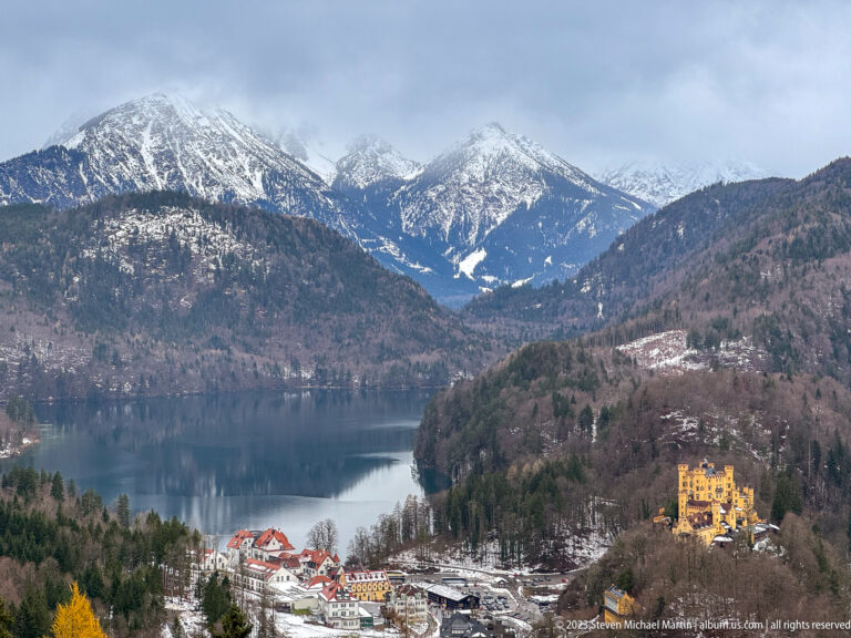 Alpsee and snow-dusted Alps with Hohenschwangau Castle on a wooded hill above the village.