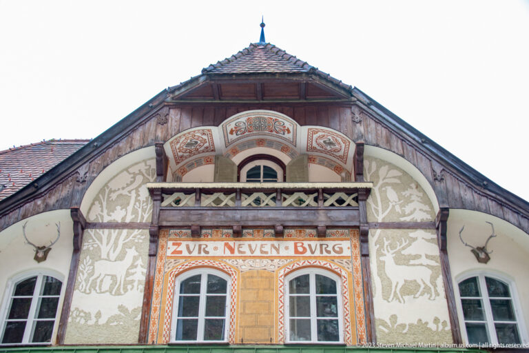 Decorated gable with painted figures, patterned borders, timber trim, and the inscription 'ZUR NEUEN BURG.'