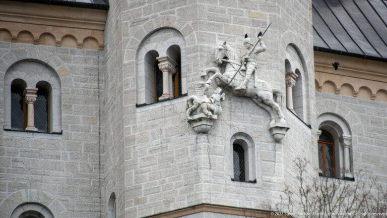 Mounted knight relief on Neuschwanstein Castle beside paired round-arched windows in pale stone masonry.
