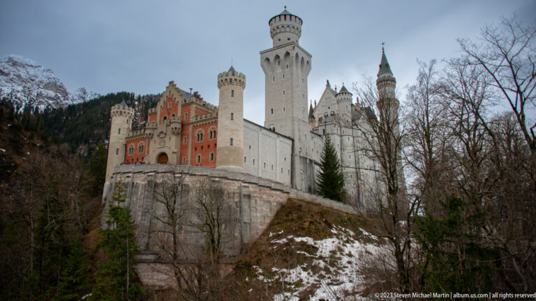 Neuschwanstein Castle above a rocky wooded slope, showing its red gatehouse, pale towers, and snowy mountains behind.
