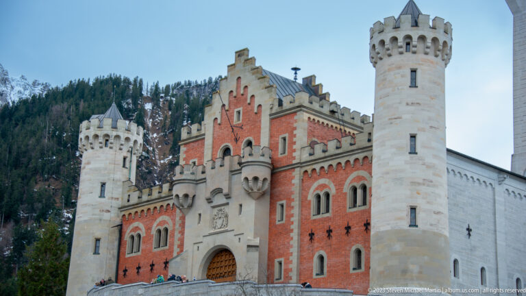 Red Neuschwanstein gatehouse between two pale cylindrical towers, with arched windows and a central entrance.