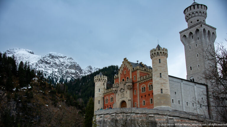 Neuschwanstein gatehouse and tall pale tower rising against snow-dusted mountains and dark winter trees.