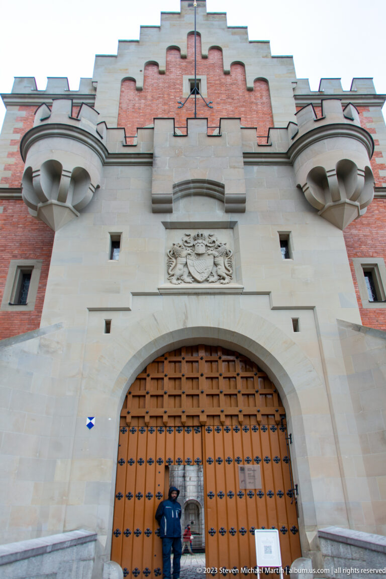 Large studded wooden gate beneath a stone arch, heraldic relief, and projecting oriels at Neuschwanstein Castle.