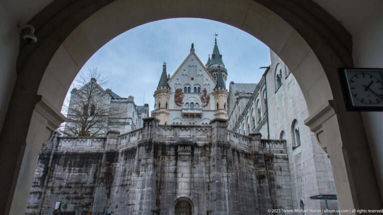 Neuschwanstein's ornate Palas façade seen through a shadowed courtyard arch, with towers, gables, and murals beyond.