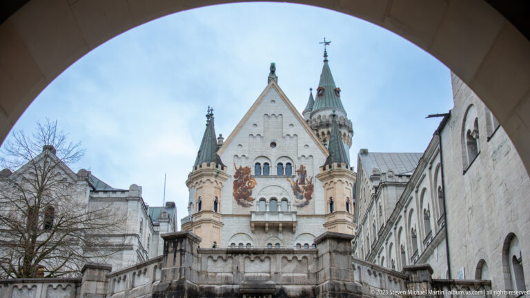 Palas façade at Neuschwanstein Castle framed by an arch, showing a steep gable, turrets, balconies, and mural decoration.