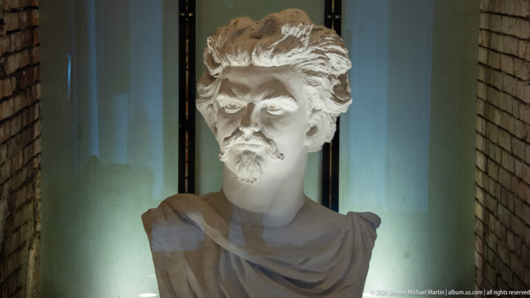 Illuminated bust of a young moustached man with swept hair and draped shoulders displayed behind glass.