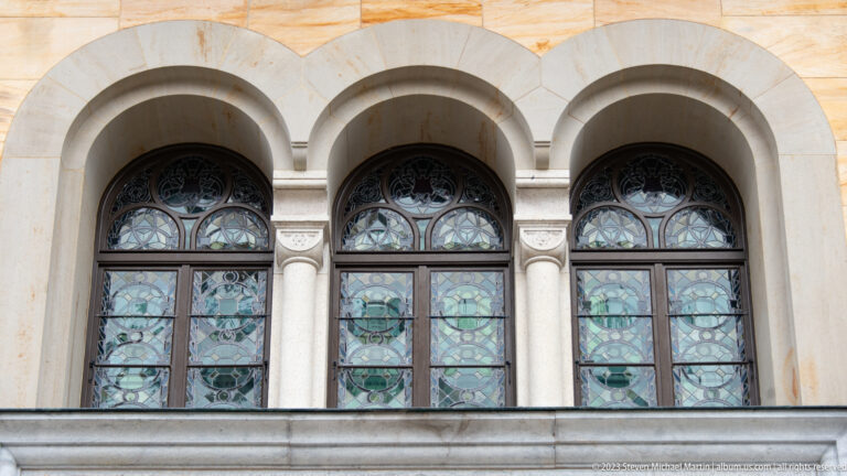Three round-arched stained-glass windows separated by slender columns in the pale stone façade of Neuschwanstein Castle.