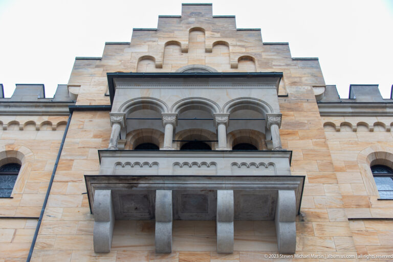 Projecting triple-arched stone balcony on a stepped pale facade at Neuschwanstein Castle, viewed from below.