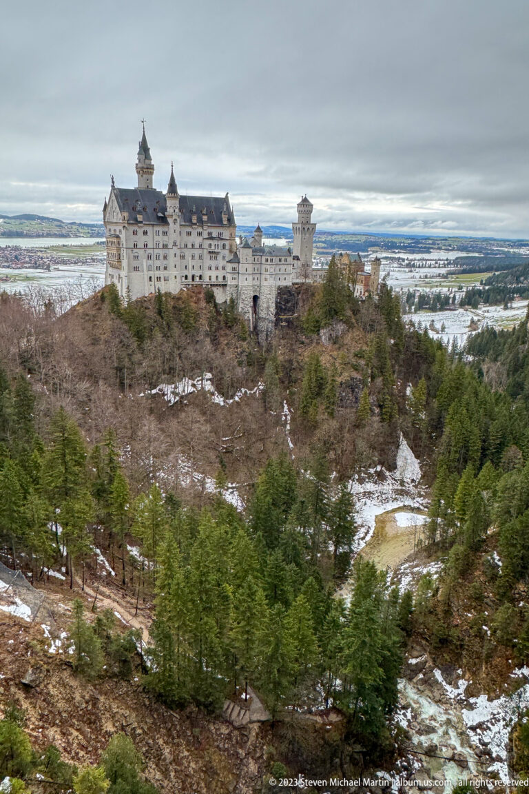 Neuschwanstein Castle across a wooded snowy ravine, with the broad Bavarian plain extending into the distance.