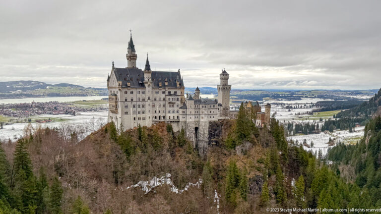 Neuschwanstein Castle seen from above across winter forest, with towers, steep roofs, and snowy fields beyond.