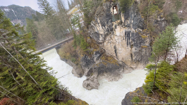 Bridge crosses a rocky river gorge beside a tall cliff with a small niche high above the water.