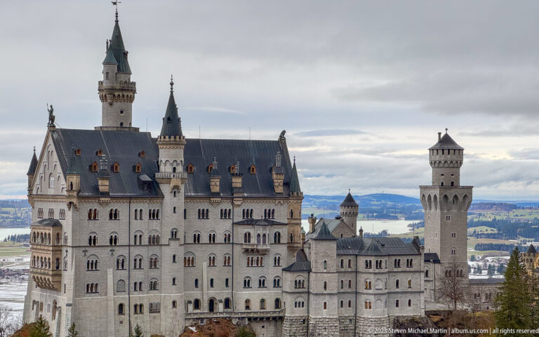 Close elevated view of Neuschwanstein Castle with pale palace walls, steep dark roofs, gables, and tall cylindrical tower.
