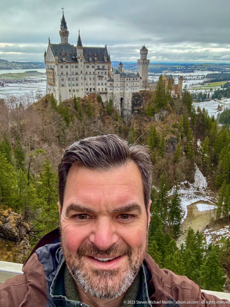 Smiling male visitor in the foreground with Neuschwanstein Castle and snowy wooded landscape behind.