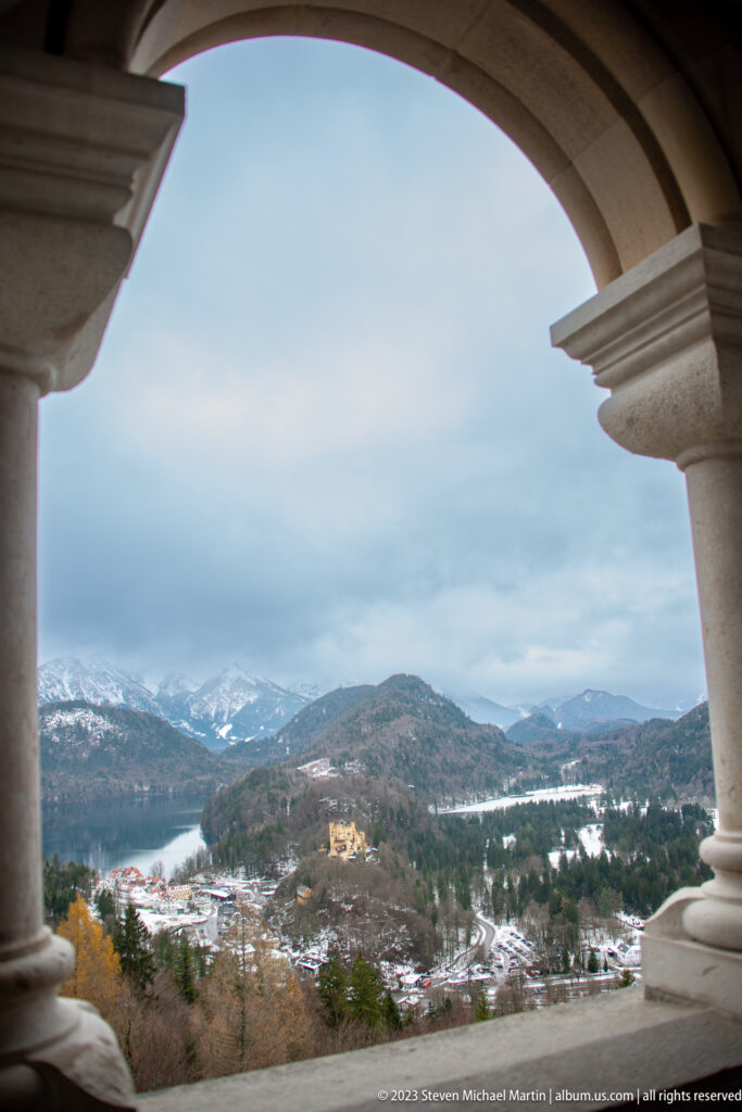 Winter valley and Hohenschwangau Castle seen through a broad stone arch and column from Neuschwanstein Castle.