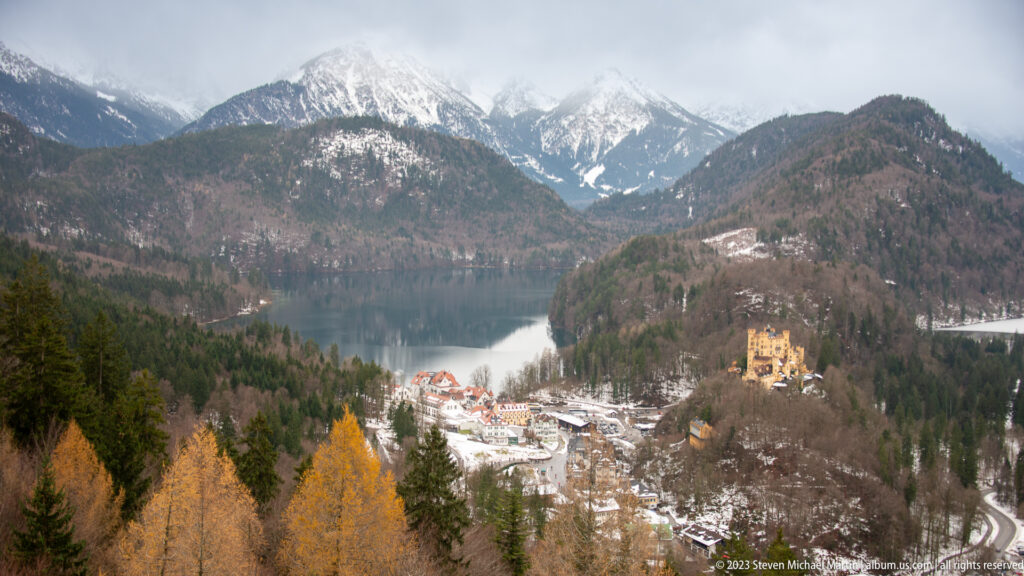 Alpsee and snowy Bavarian Alps above Hohenschwangau, with the ochre castle visible on a wooded hill.